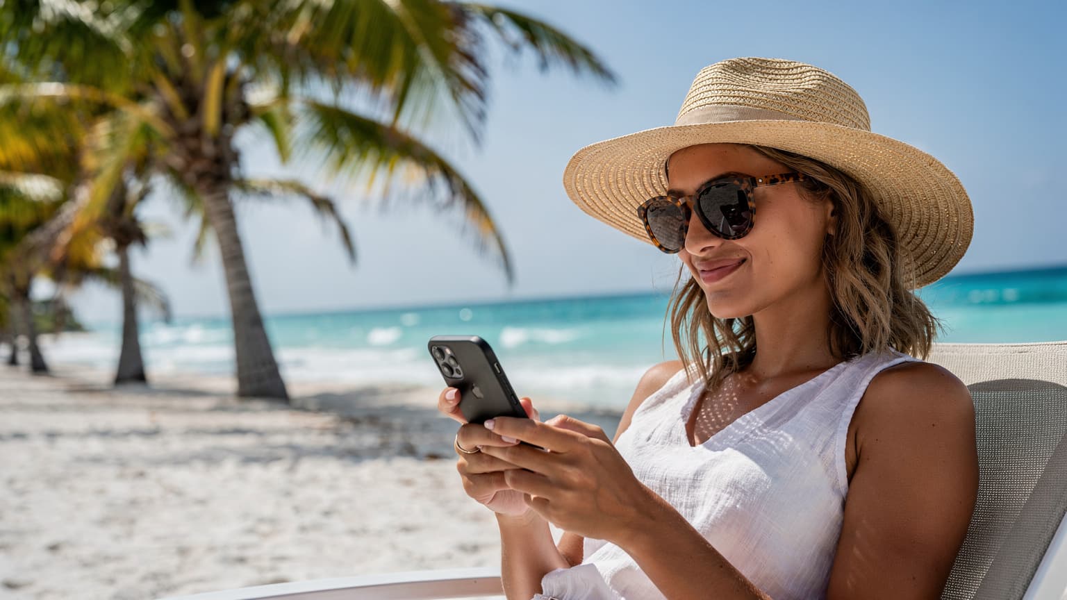 Una mujer joven con gafas de sol y sombrero de ala ancha mirando su teléfono en una playa soleada de Cancún, con palmeras y el mar Caribe de fondo.