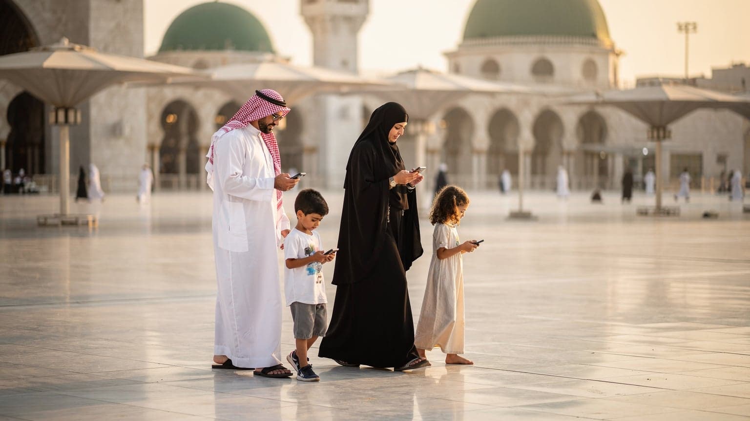 عائلة سعودية، أب وأم وطفلان، ينظرون إلى هواتفهم الذكية وهم يسيرون في ساحة المسجد النبوي بالمدينة المنورة في ضوء المساء الدافئ، يظهر في الخلفية جزء من المسجد.