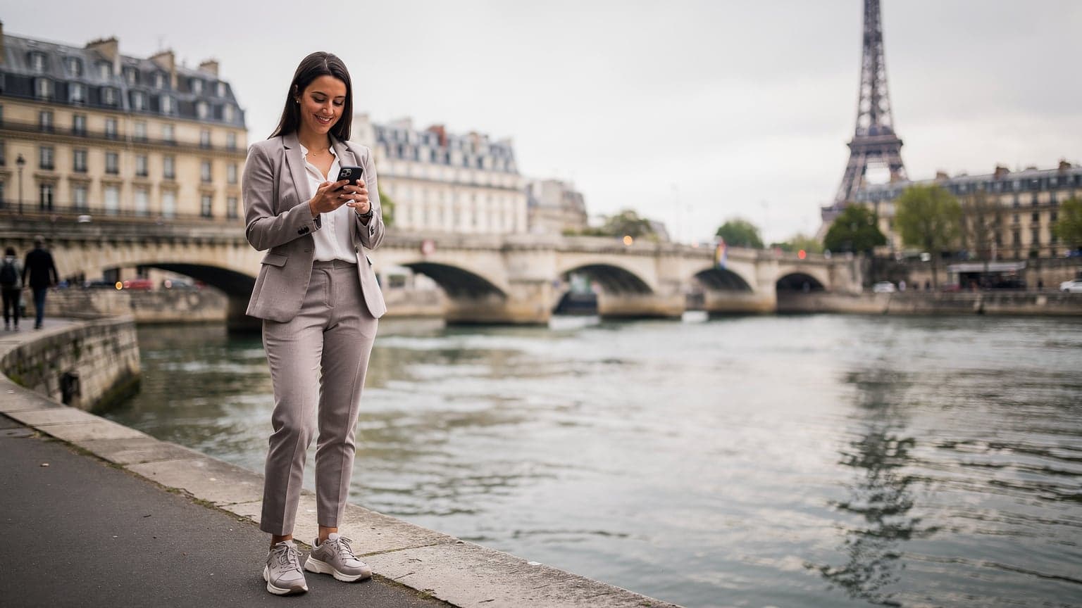 Une femme souriante, téléphone en main, naviguant sur les quais de Seine avec la Tour Eiffel en arrière-plan, sous un ciel légèrement couvert.
