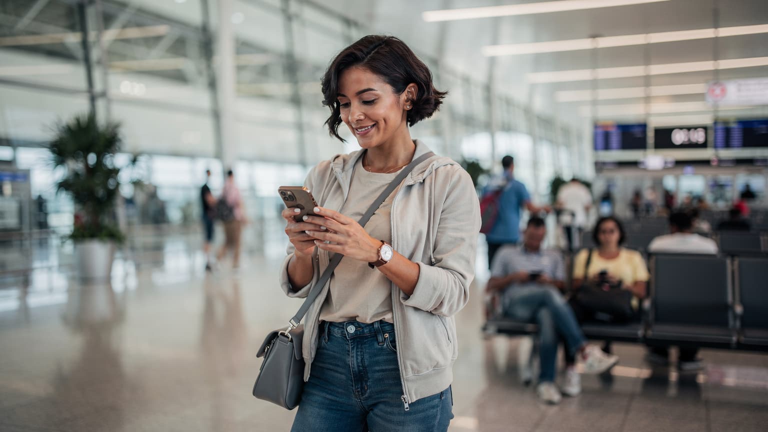 Seorang wanita tersenyum sambil memegang ponsel di Bandara Changi Singapura, dengan latar belakang gerbang keberangkatan yang ramai.