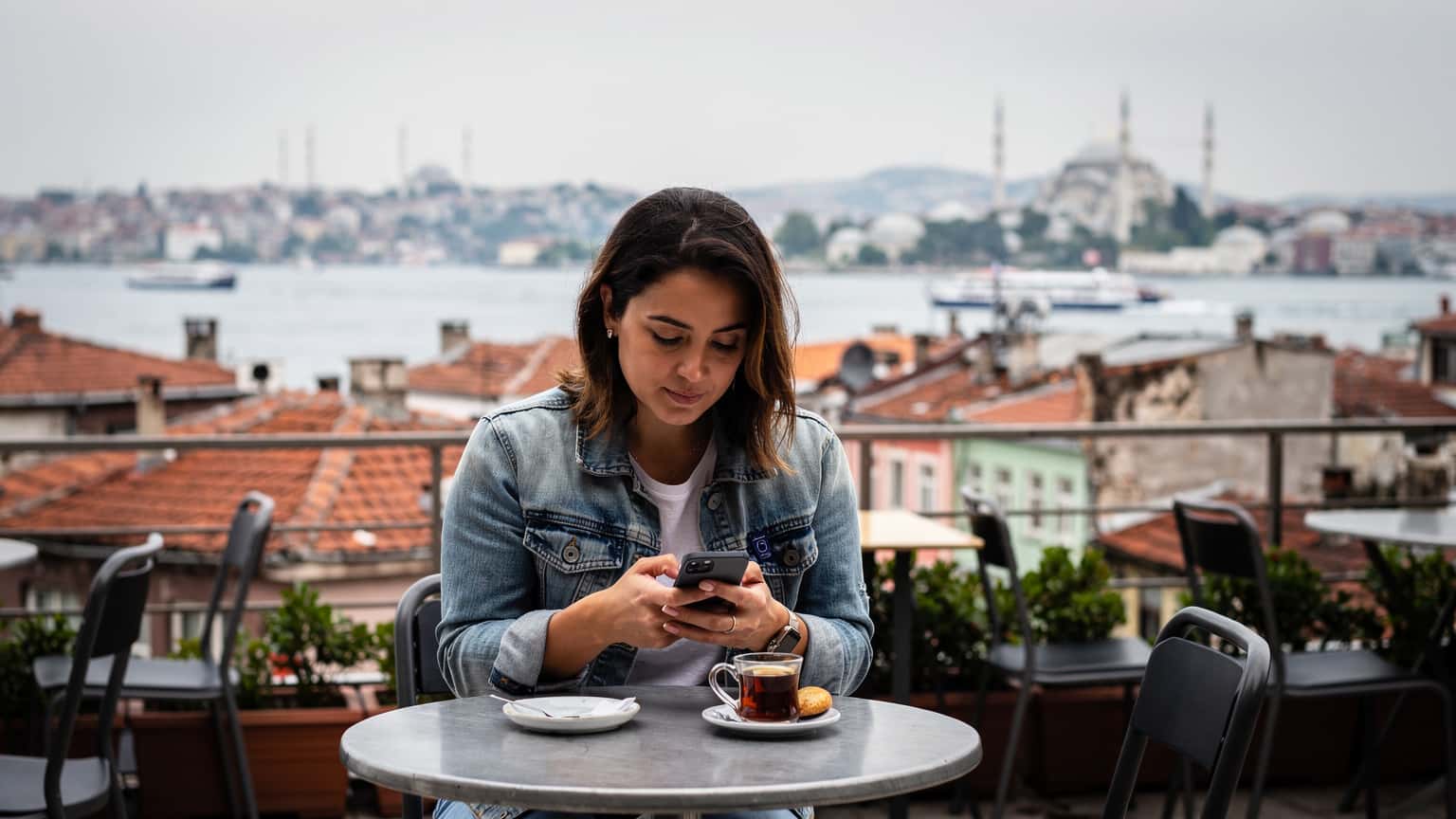 Une femme aux cheveux bruns, vêtue d'une veste en jean, consulte son téléphone avec un air concentré sur la terrasse d'un café traditionnel à Istanbul, avec le Bosphore et des toits rouges en arrière-plan sous un ciel légèrement nuageux.