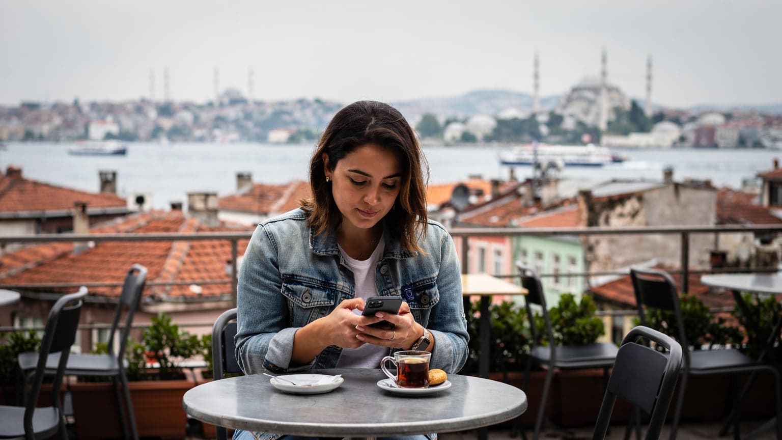 Une femme aux cheveux bruns, vêtue d'une veste en jean, consulte son téléphone avec un air concentré sur la terrasse d'un café traditionnel à Istanbul, avec le Bosphore et des toits rouges en arrière-plan sous un ciel légèrement nuageux.
