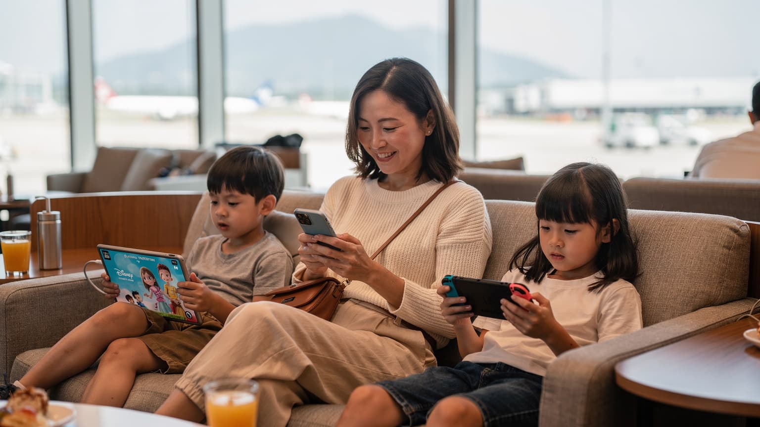 一位母亲在香港国际机场候机室里, 她的两个孩子分别坐在旁边看iPad和玩Nintendo Switch, 母亲正微笑着在自己的iPhone上查看eSIM设置。