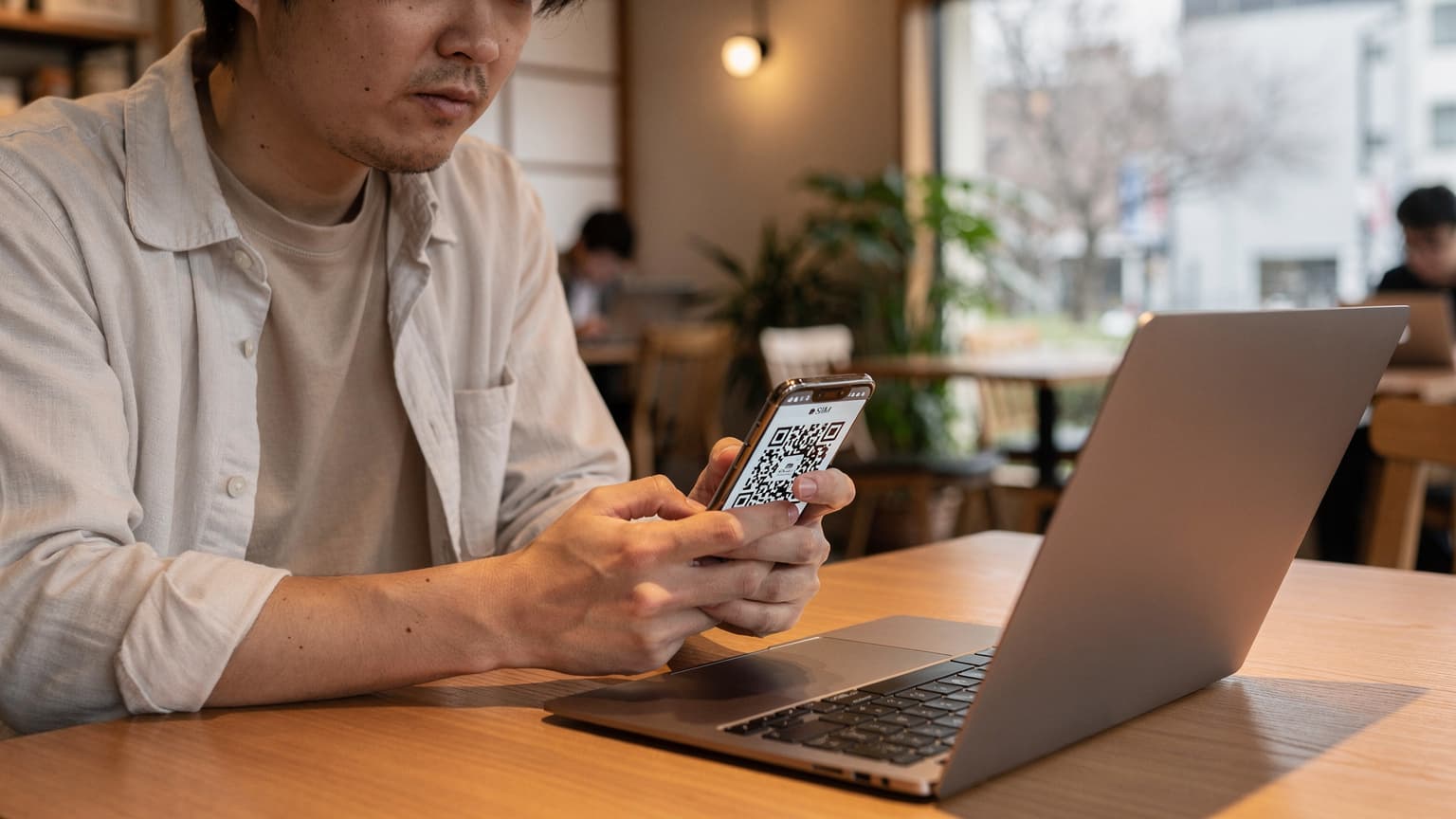 東京の新宿御苑近くのカフェで、ノートPCとスマートフォンを広げ、eSIMの設定画面を確認する旅行者の手元。窓からは都会のビル群が見える。