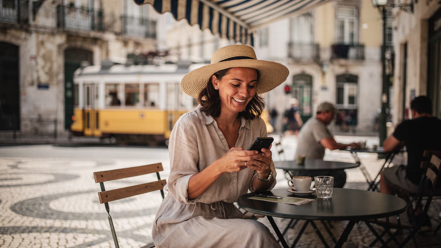 Uma mulher sorridente com um chapéu de sol, usando seu smartphone para verificar um mapa digital enquanto se senta em um café charmoso no bairro da Baixa, em Lisboa, com um elétrico amarelo passando ao fundo.