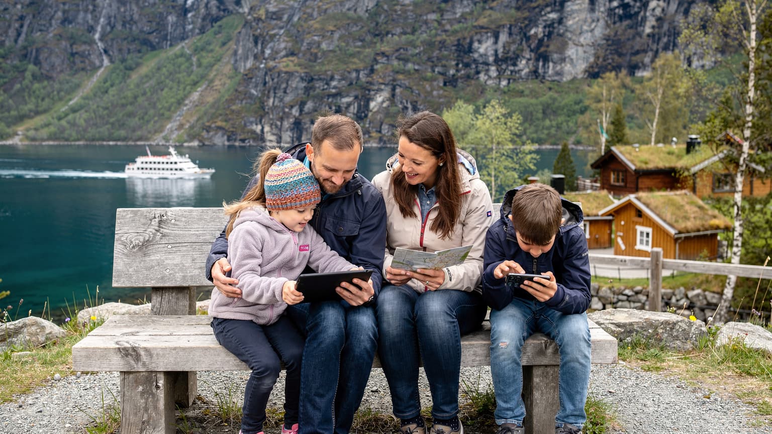 En familie på fire, to voksne og to barn i alderen 8 og 10, sitter på en benk ved en innsjø i de norske fjellene, med mobiltelefoner og nettbrett i bruk, omgitt av frodig grønt og snødekte topper i bakgrunnen.