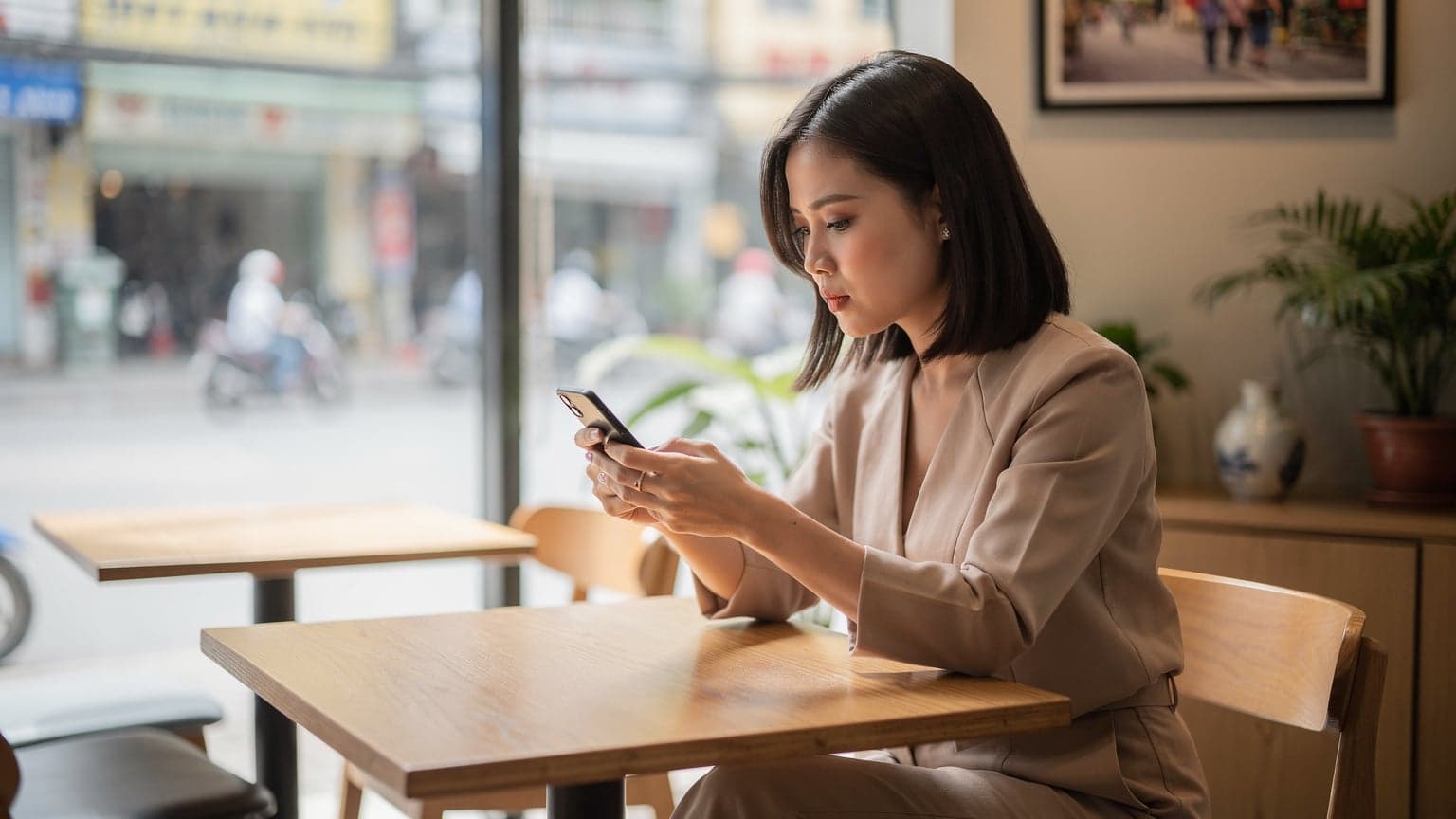 Một người phụ nữ trẻ đang ngồi tại một quán cà phê hiện đại ở Thành phố Hồ Chí Minh, chăm chú nhìn vào màn hình điện thoại thông minh của mình, với một ly cà phê đá và máy tính xách tay mở trên bàn.
