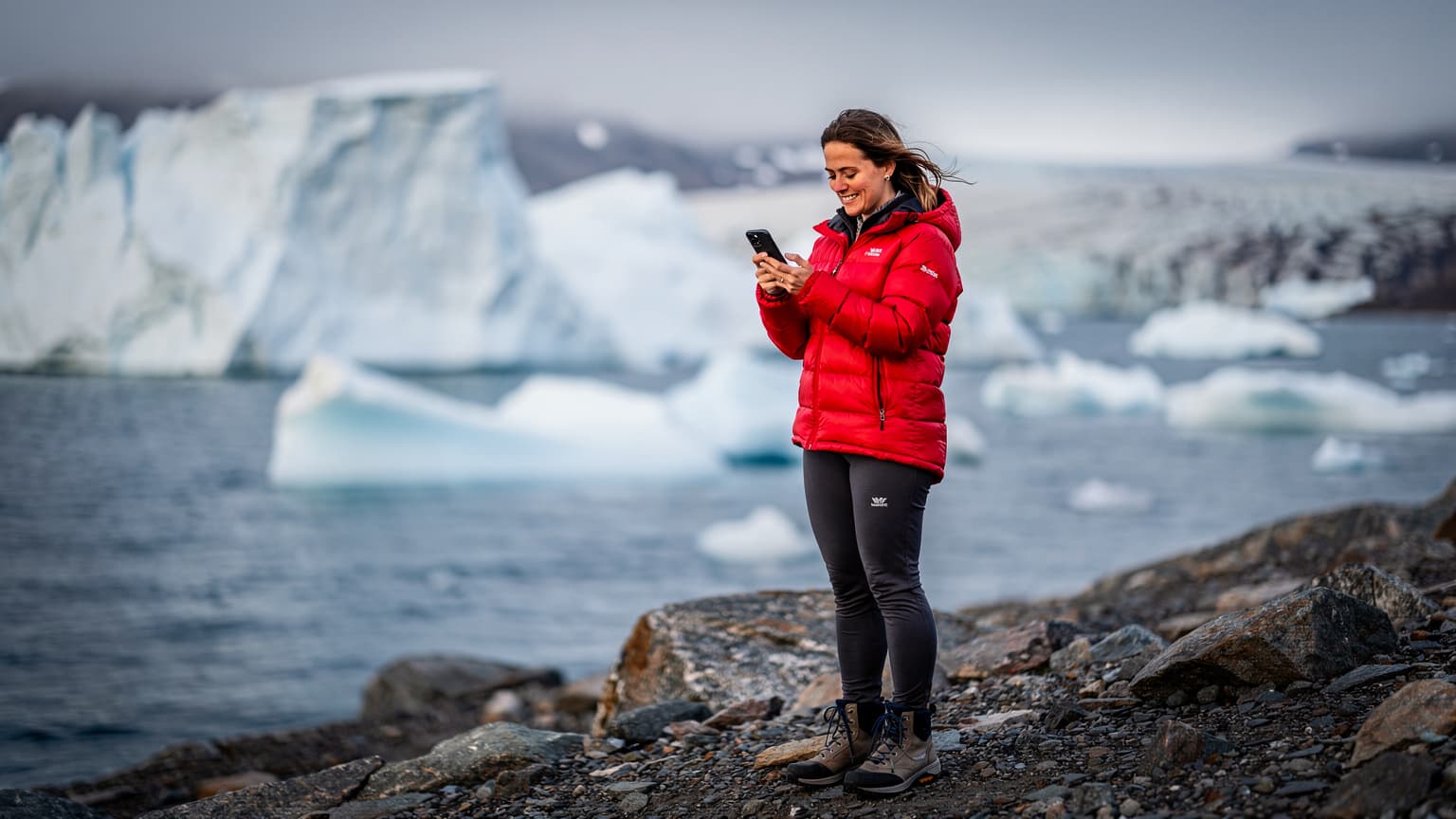 En kvinde i en varm dunjakke står på en kystlinje i Ilulissat, Grønland, med isbjerge i baggrunden, kigger på sin telefon med et smil.