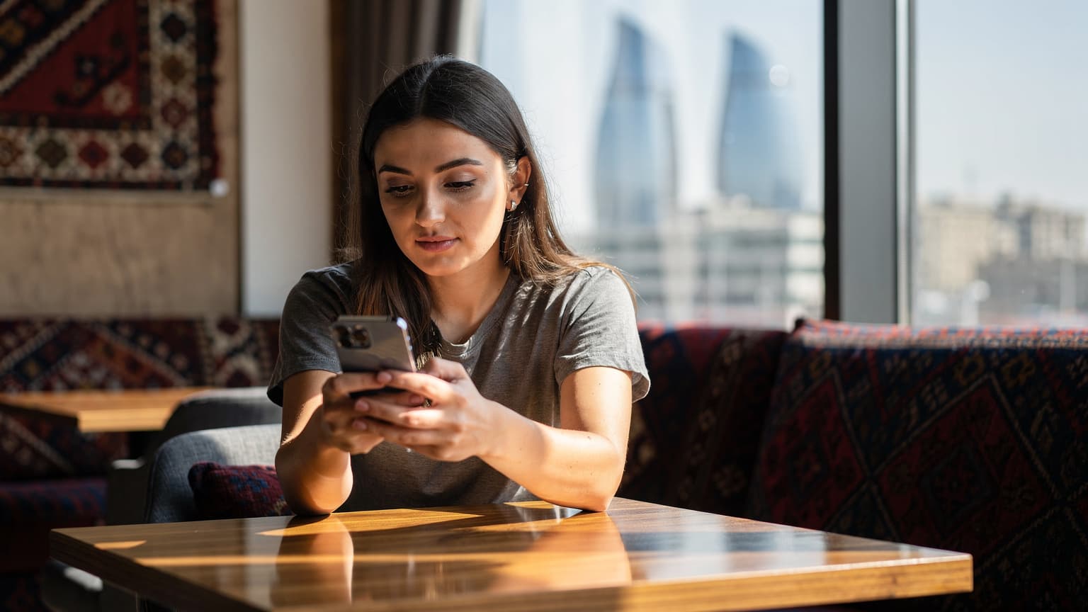 Eine junge Frau sitzt an einem Tisch in einem modernen Café in Baku, Aserbaidschan, und schaut auf ihr Smartphone, während sie eine Tasse Tee trinkt.