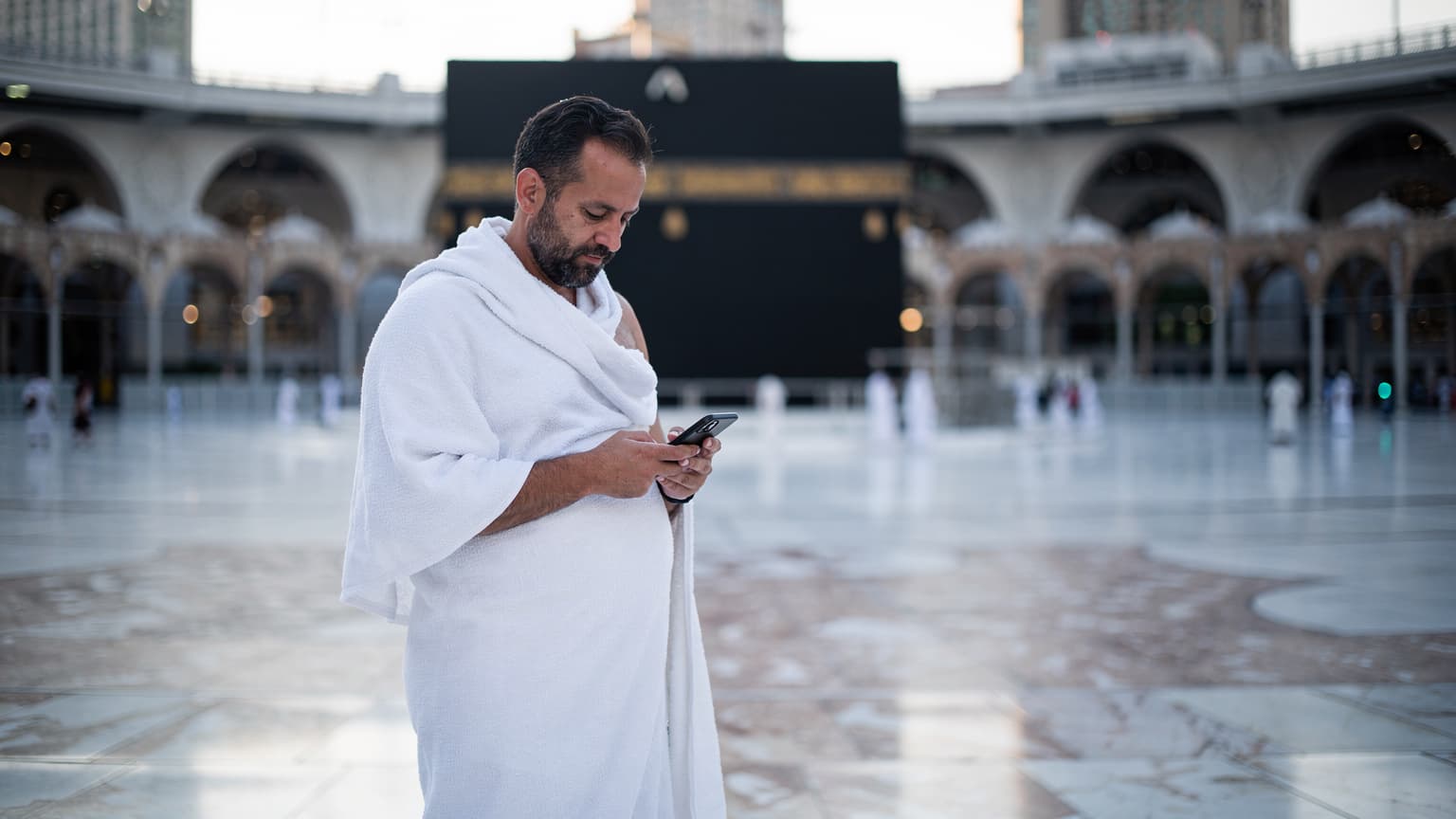 حاج يحمل هاتفه الذكي ويفحص خريطة في صحن الكعبة المشرفة في مكة المكرمة، مع إضاءة خفيفة وقت الفجر تظهر تفاصيل العمارة الإسلامية