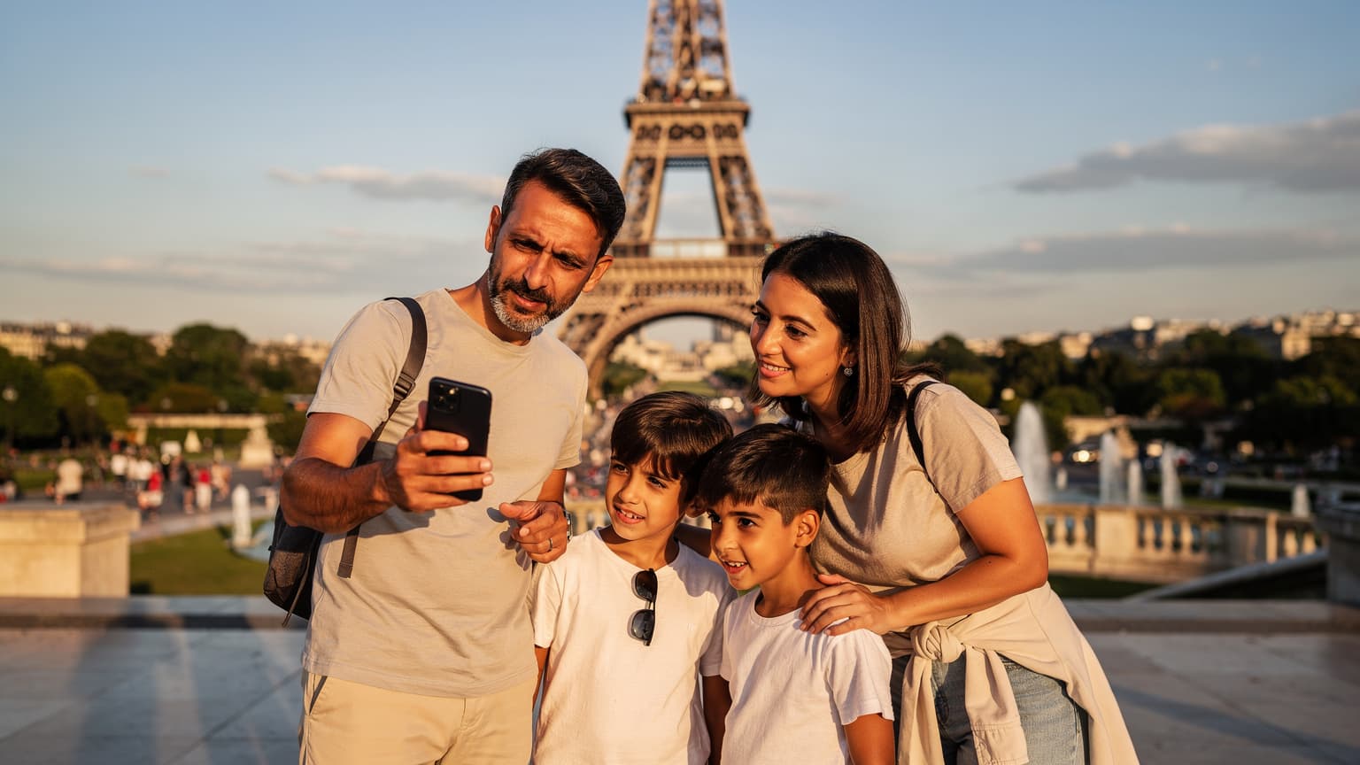 Paris'te Eyfel Kulesi önünde, ailece telefonlarından harita kontrol eden mutlu bir Türk ailesi, arka planda güneş batımı.