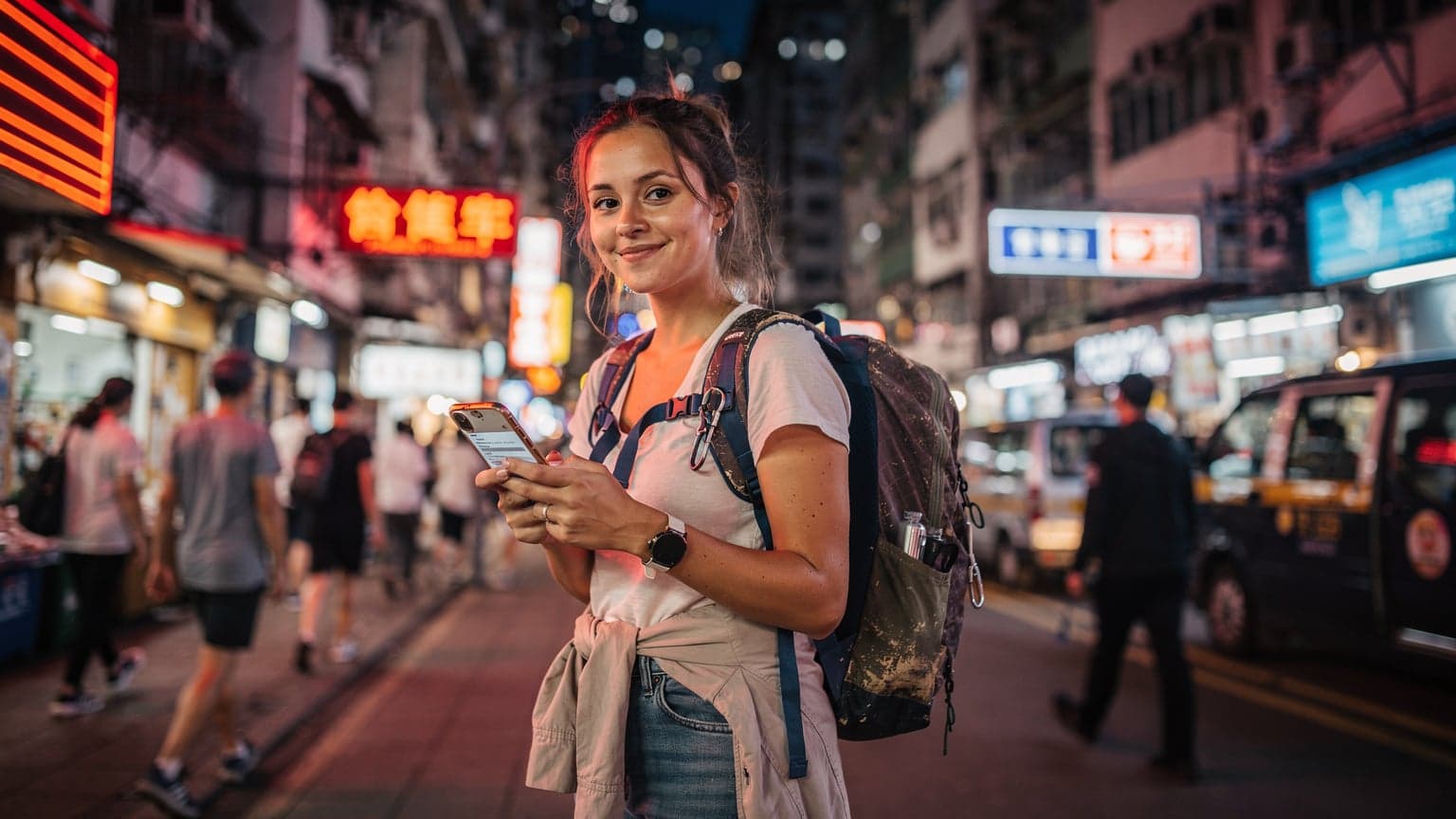 一位年轻的旅行者在香港旺角的街头,手持一部显示 eSIM 设置界面的 iPhone,背景是霓虹灯闪烁的招牌和穿梭的人群,脸上带着轻松愉快的笑容。