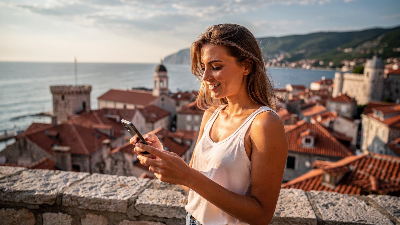 Mladá žena se smartphonem v ruce, usmívá se a sleduje něco na obrazovce, s tyrkysovým mořem a střechami starého města Budvy v pozadí za slunečného odpoledne.