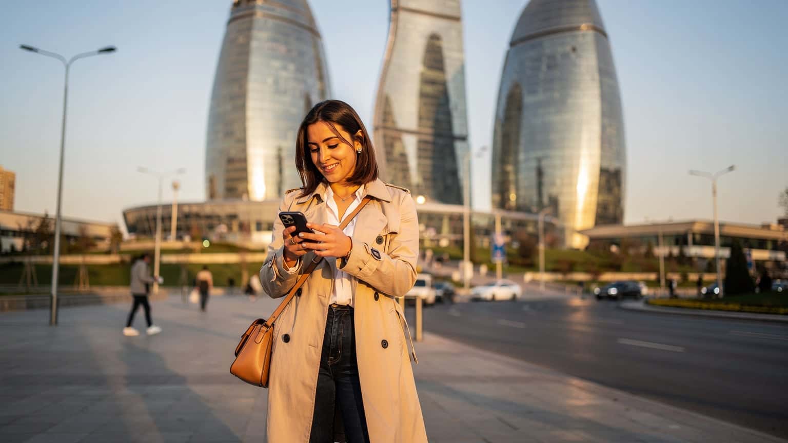 Bakü'deki Flame Towers'ın önünde, telefonunda harita uygulamasına bakan gülümseyen bir kadın.