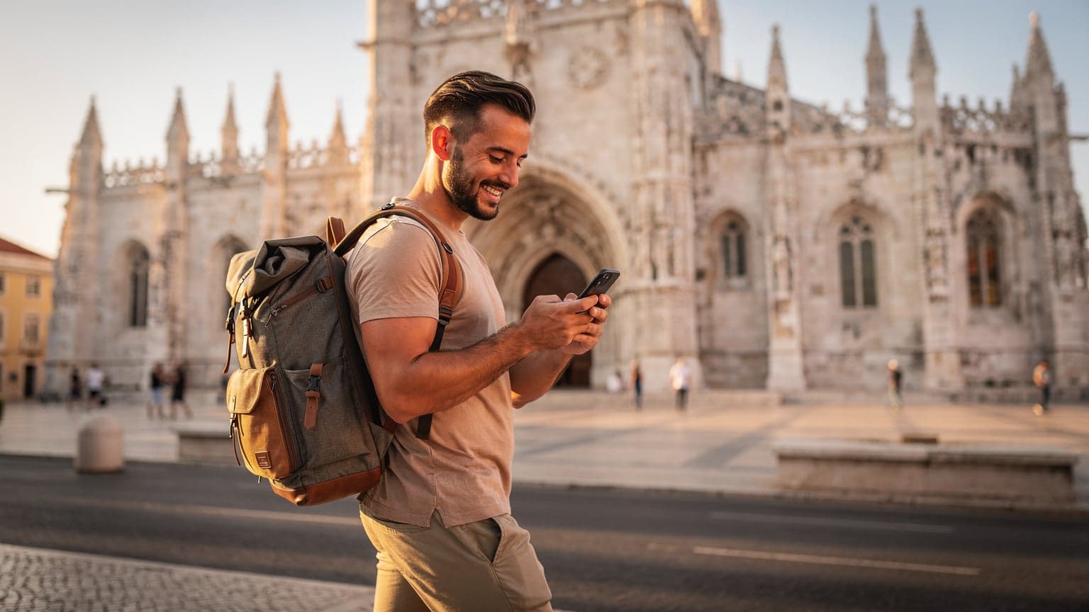 Um viajante olhando o Mosteiro dos Jerónimos em Lisboa, verificando o telefone para conectividade com um sorriso satisfeito, com a luz dourada do final da tarde iluminando a cena.