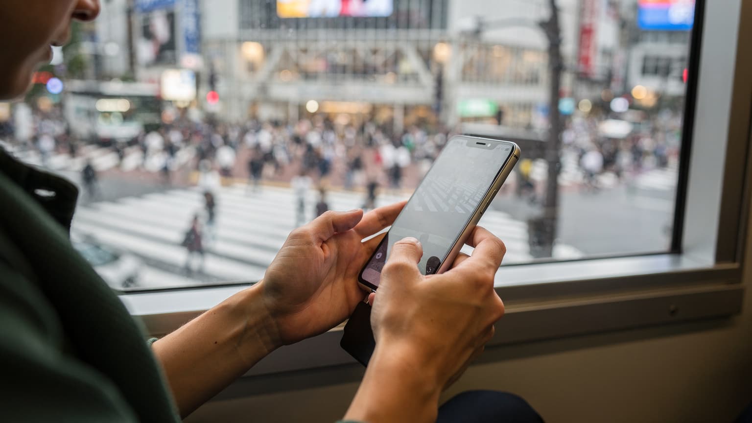 東京の山手線車内で、窓の外の渋谷スクランブル交差点を眺めながらスマートフォンを操作する日本人旅行者の手元がクローズアップされている。