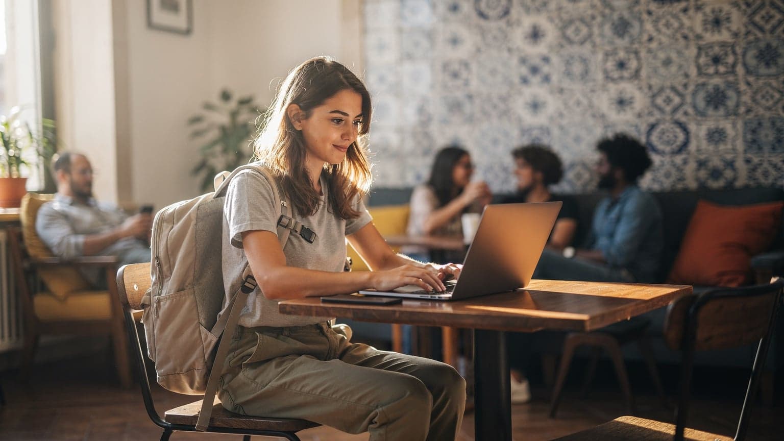 A young female backpacker in a colorful hostel common room in Lisbon, Portugal, typing on a modern laptop with an active eSIM connection, a half-eaten pastry and a small coffee cup on the table next to her.