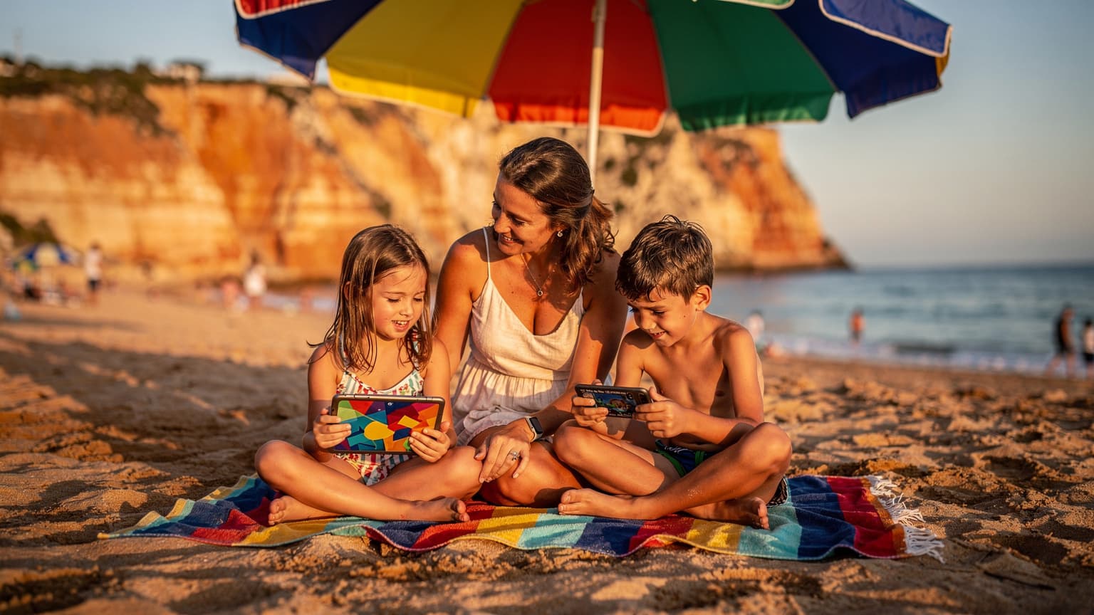 Uma mãe sorridente e seus dois filhos, um com um tablet e outro com um smartphone, sentados em uma toalha de praia colorida sob um guarda-sol na Praia da Falésia, no Algarve, Portugal.