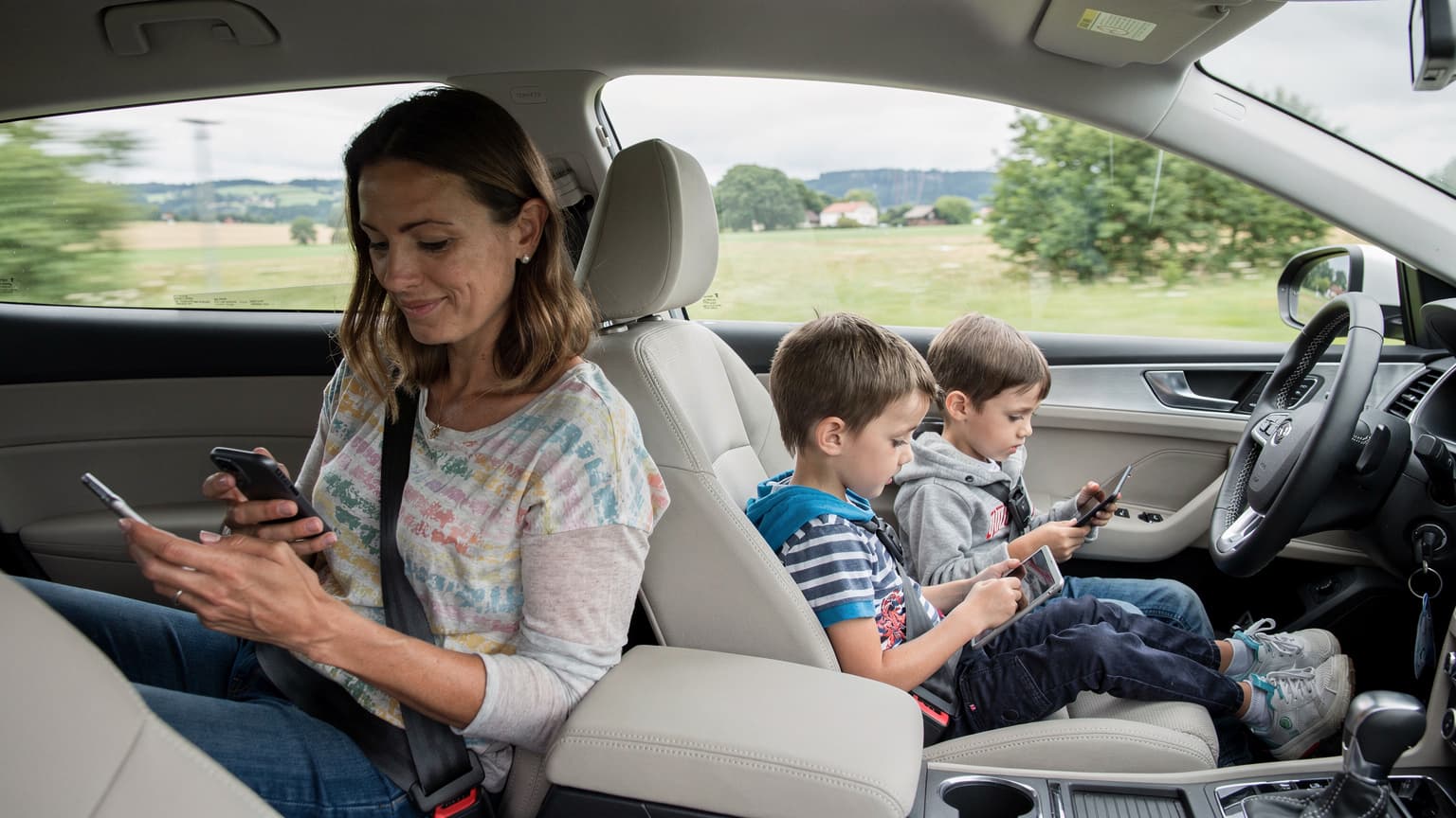 A mother calmly looking at her smartphone while her two children, aged around 7 and 10, are engrossed in their tablets in the back seat of a car on a long road trip.
