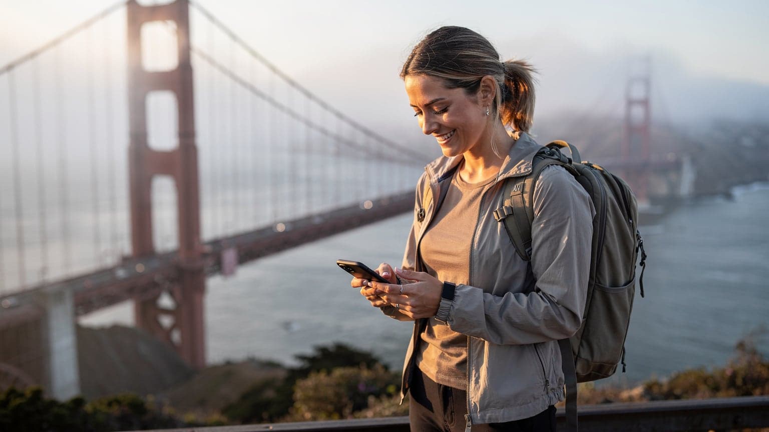 A woman looks at her phone, smiling, while standing in front of the iconic Golden Gate Bridge in San Francisco, with the morning fog lifting.
