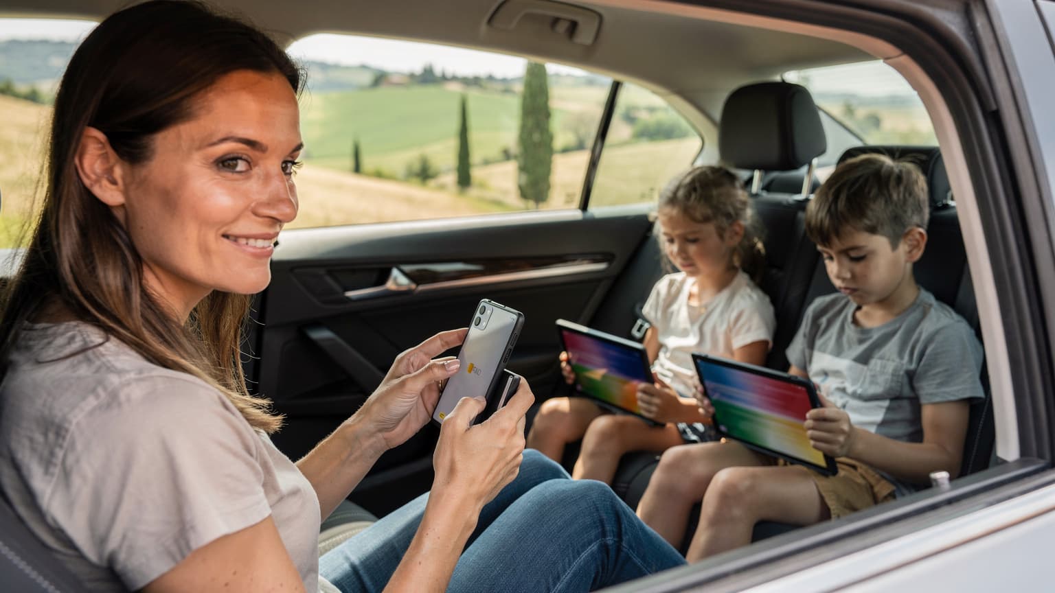A mother calmly setting up eSIM on her smartphone while her two children, aged 6 and 10, are engrossed in their tablets in the back seat of a rental car, driving through the scenic Italian countryside near Tuscany.