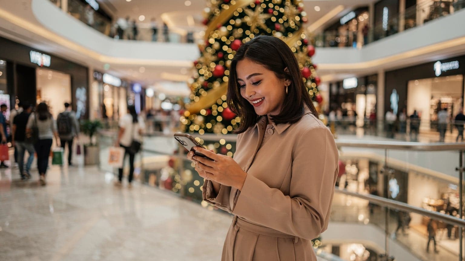 Seorang wanita tersenyum sambil memegang ponselnya di depan pohon Natal yang dihias indah di pusat perbelanjaan Grand Indonesia Jakarta, dengan banyak orang berbelanja di latar belakang.