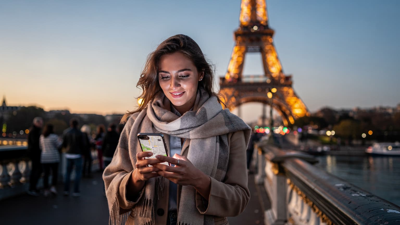 Une femme souriante, téléphone en main, naviguant sur une carte numérique près de la Tour Eiffel illuminée au crépuscule, avec une foule animée en arrière-plan.