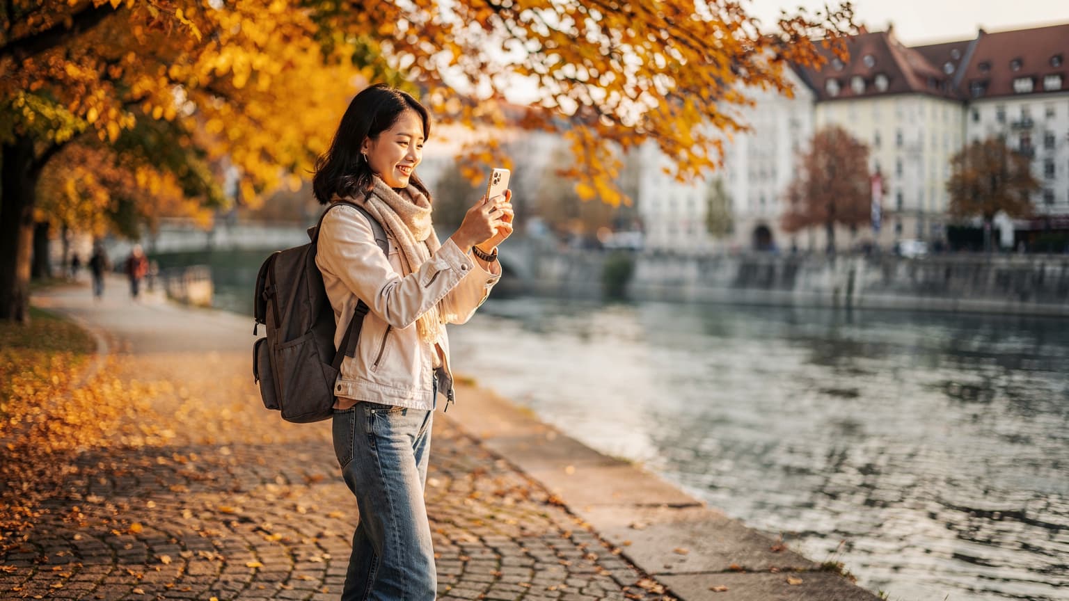 Một phụ nữ Việt Nam trẻ tuổi, đeo ba lô, đang mỉm cười và giơ điện thoại lên chụp ảnh những hàng cây phong lá vàng rực rỡ bên bờ sông Isar ở Munich, Đức, với một tòa nhà cổ kính thấp thoáng phía sau.