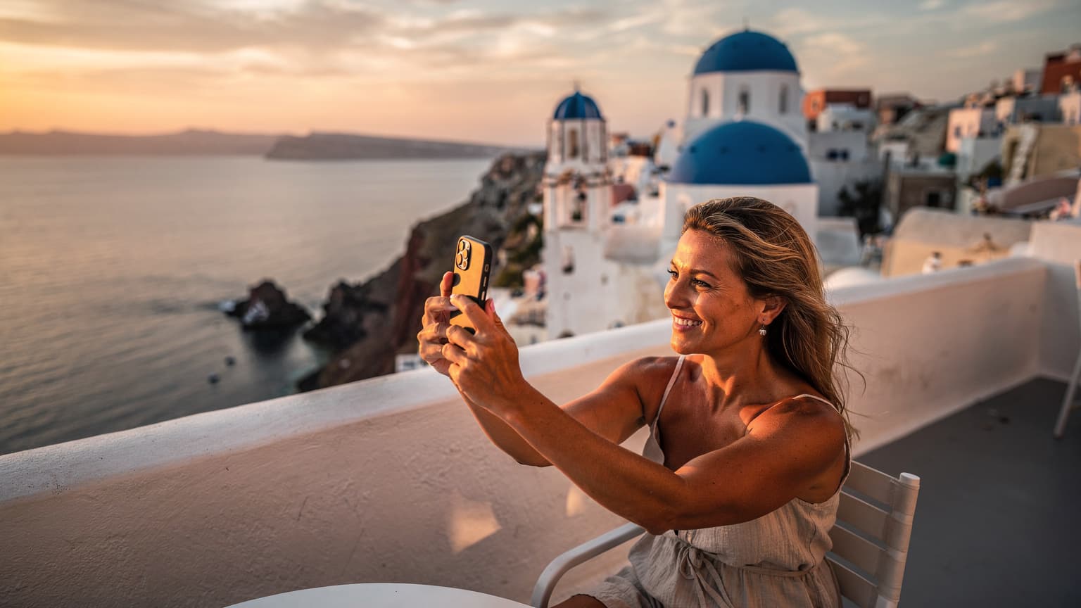 En kvinna sitter på en vitkalkad terrass i Oia, Santorini, med utsikt över Egeiska havet och solnedgången, leende medan hon håller sin smartphone.