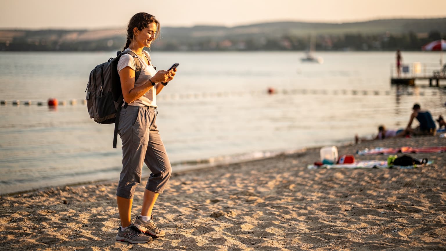Mladá žena s batohom pozerá na mobilný telefón s úsmevom na pláži jazera Balaton v Maďarsku pri západe slnka.