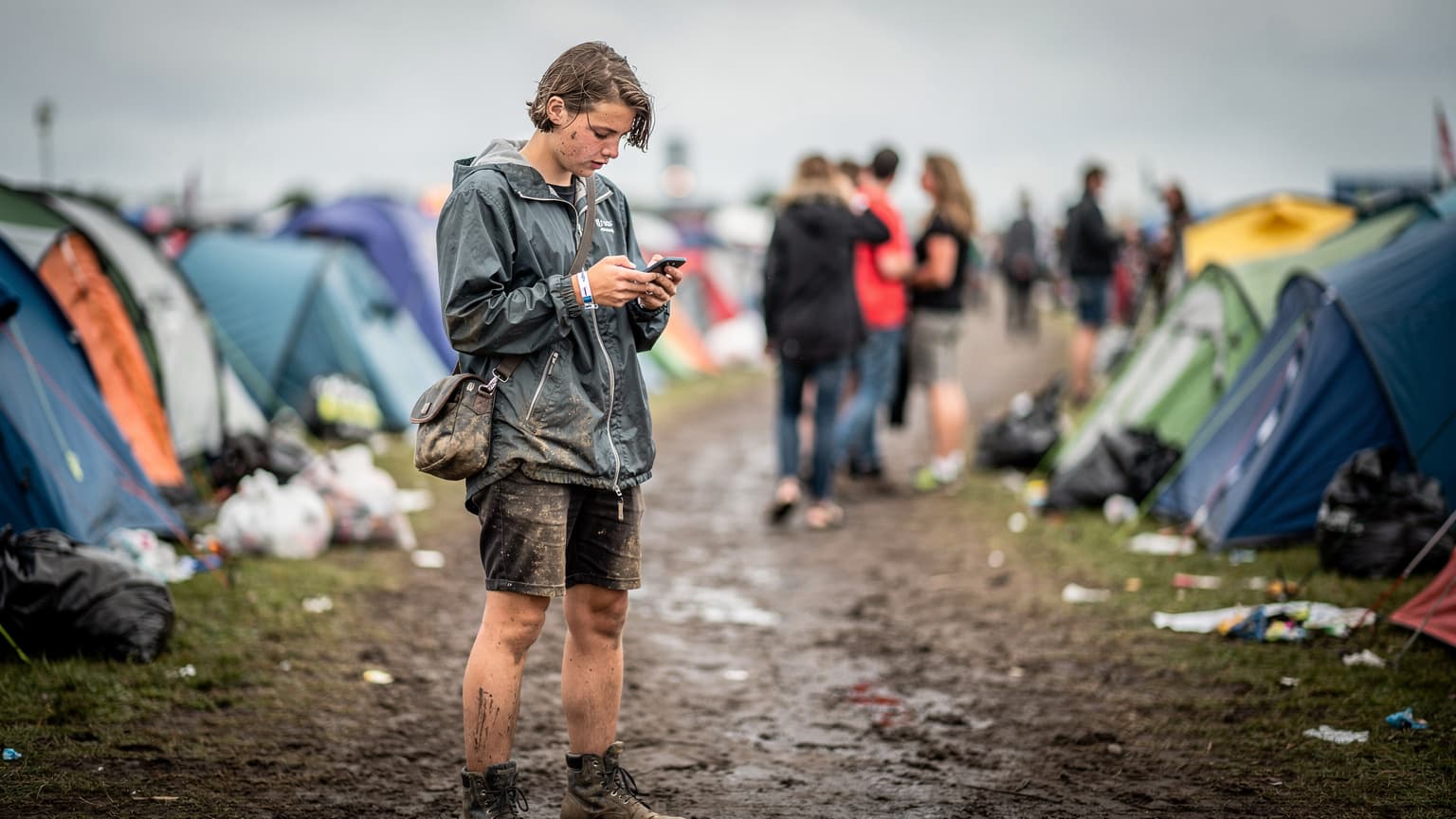 En festivalgænger tjekker sin telefon for at finde vej gennem en mudret sti på Roskilde Festival, omgivet af telte og glade mennesker.