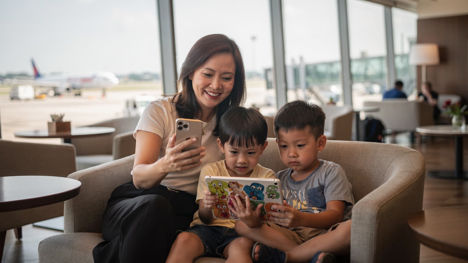 Một người phụ nữ Việt Nam đang mỉm cười, cầm điện thoại iPhone trong tay, cùng với hai đứa trẻ đang xem hoạt hình trên iPad tại sảnh chờ sân bay quốc tế Changi, Singapore, với ánh sáng tự nhiên mềm mại chiếu qua cửa sổ lớn.