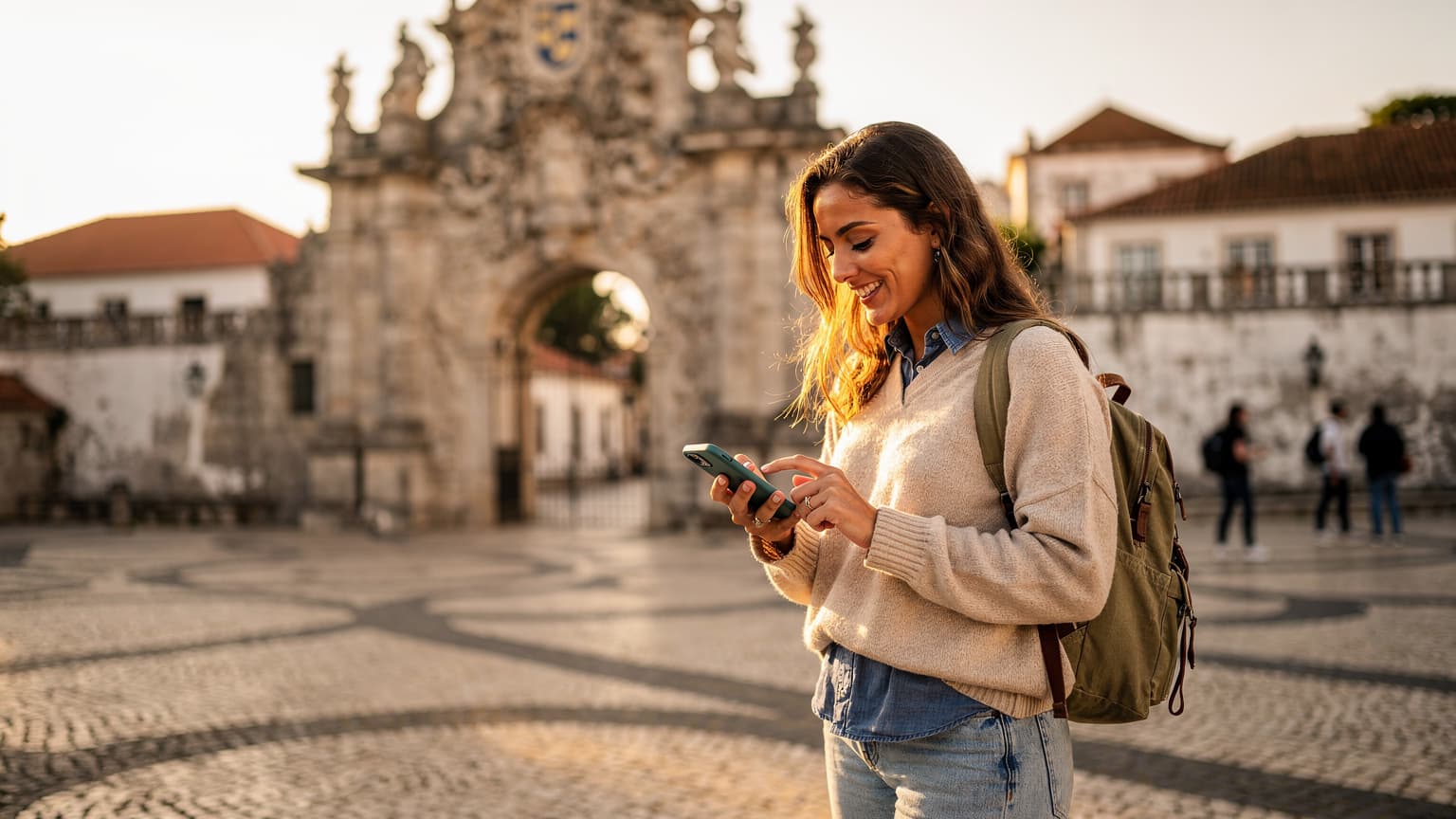 Uma estudante sorridente, com mochila, ajustando seu celular com eSIM em frente à histórica Universidade de Coimbra, sob um céu de fim de tarde de setembro.