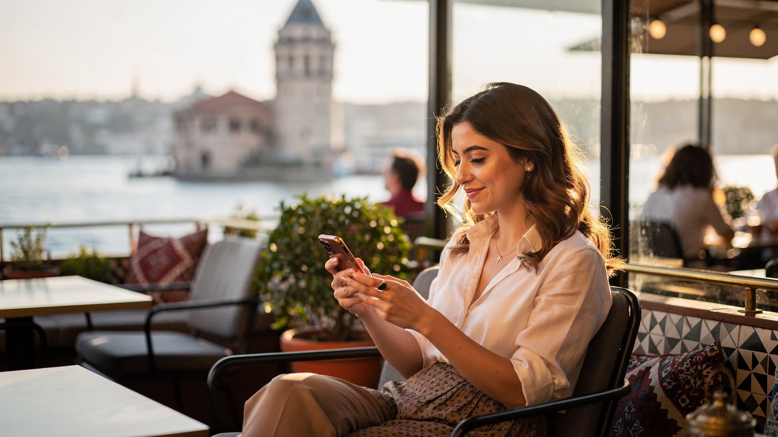 İstanbul'un Karaköy semtinde, galata kulesi manzaralı bir kafede oturan, elinde akıllı telefonuyla eSIM kurulumu yapan genç bir kadın.