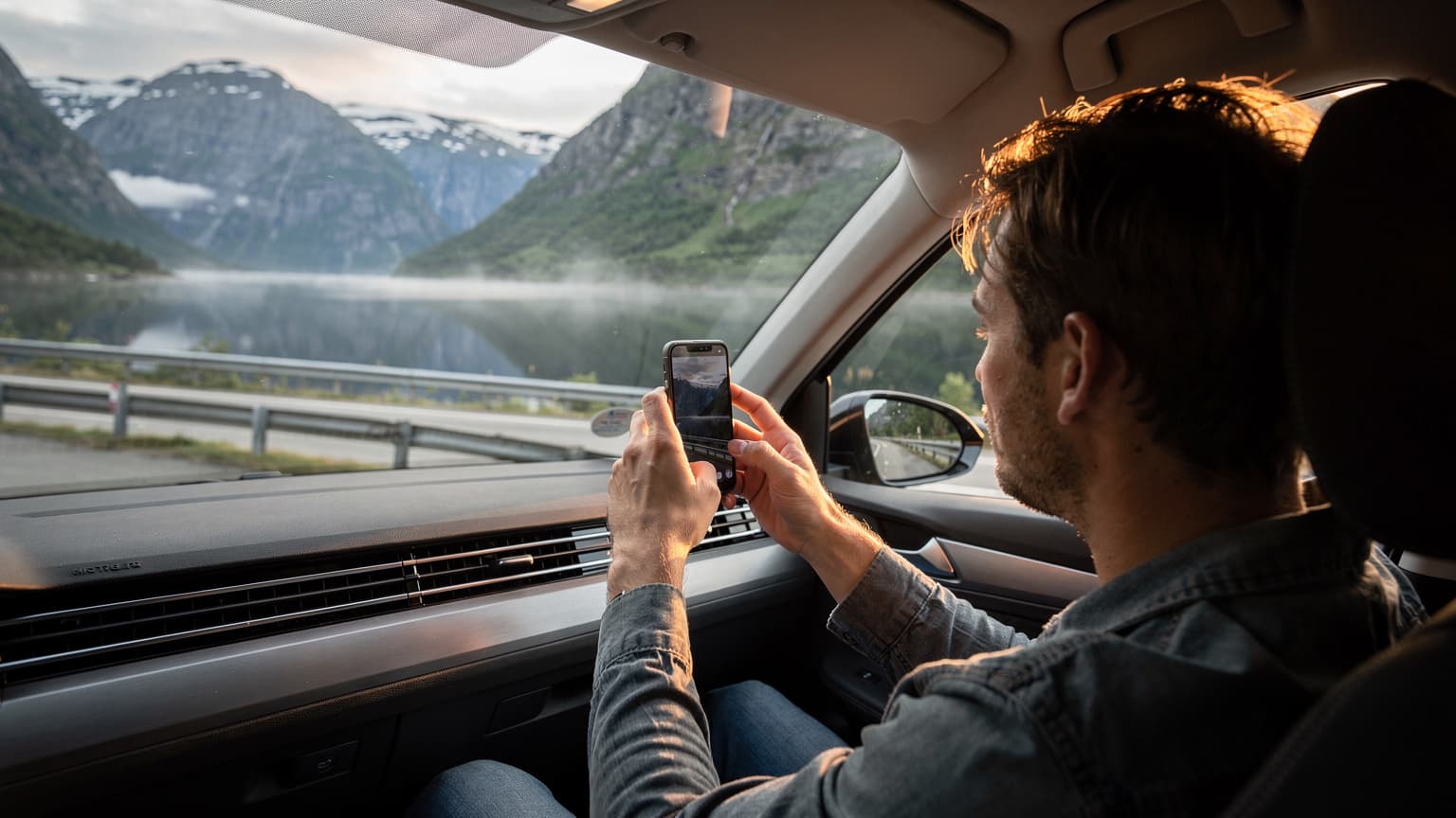 En bil kör längs en slingrande väg i norska fjordlandskapet med en person som håller upp en smartphone för att fånga utsikten, i mjukt morgonljus.