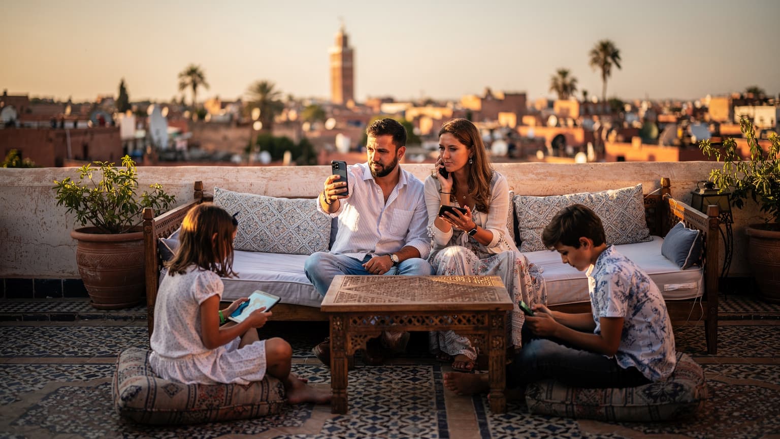 Une famille française, parents et deux enfants, utilise leurs téléphones et tablettes sur la terrasse d'un riad traditionnel à Marrakech, avec des palmiers et la Koutoubia en arrière-plan, au coucher du soleil.