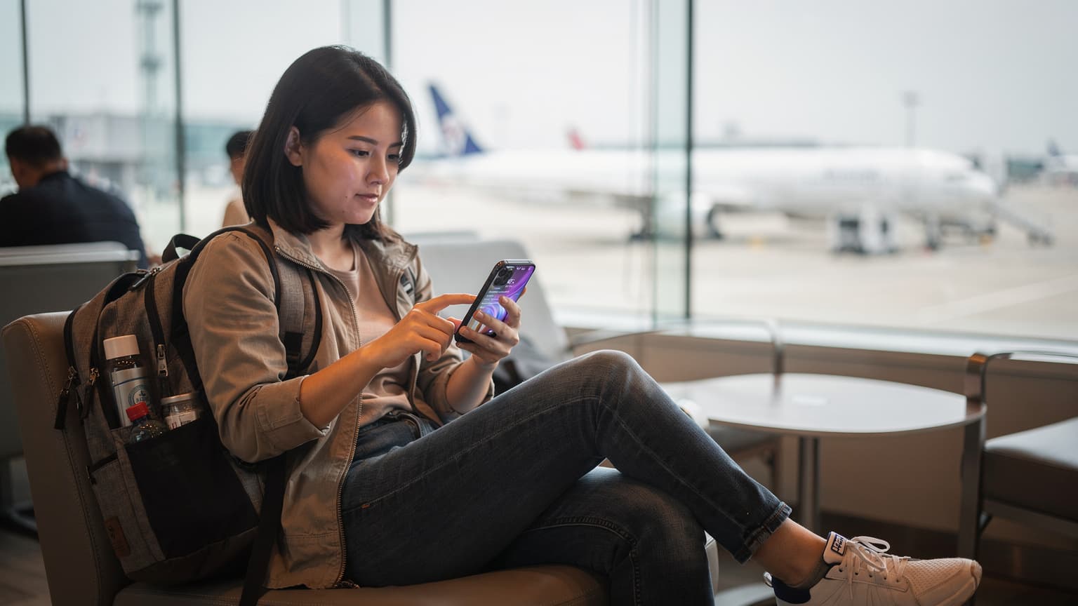 一名香港旅客在機場候機室內,手持一台 iPhone,屏幕上顯示著 eSIM 設置界面,窗外是繁忙的停機坪景色。
