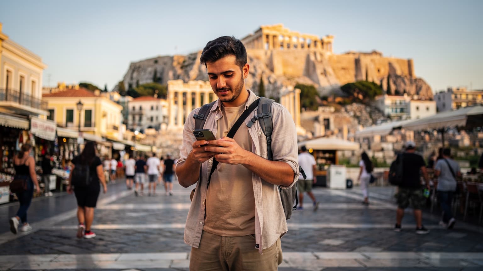 Ένας νεαρός άνδρας χρησιμοποιεί το smartphone του, στέκεται σε μια πολυσύχναστη πλατεία στην Αθήνα, με φόντο την Ακρόπολη στο ηλιοβασίλεμα.