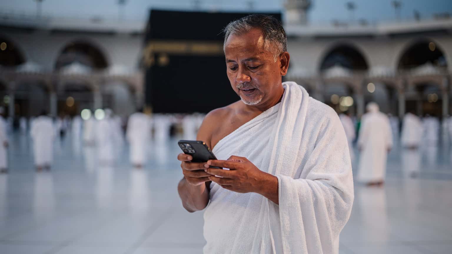 Seorang jemaah Malaysia berumur 50-an, memakai ihram, sedang melihat skrin telefonnya dengan tenang di perkarangan Masjidil Haram, Mekah, pada waktu subuh.