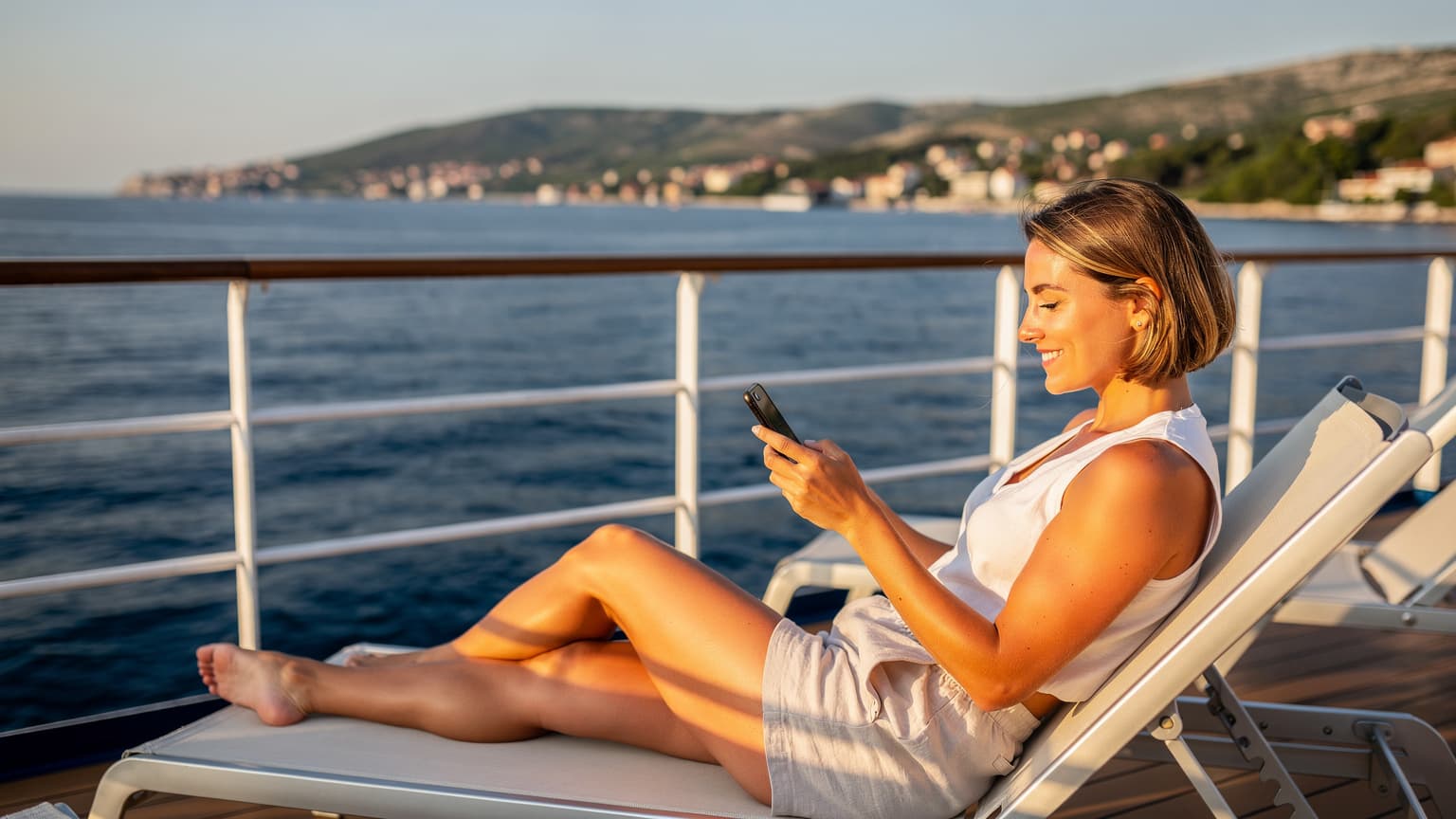 Eine junge Frau lächelt entspannt, während sie auf dem Deck eines Kreuzfahrtschiffes in der Adria sitzt und auf ihr Smartphone schaut, mit dem Meer und der Küste im Hintergrund.