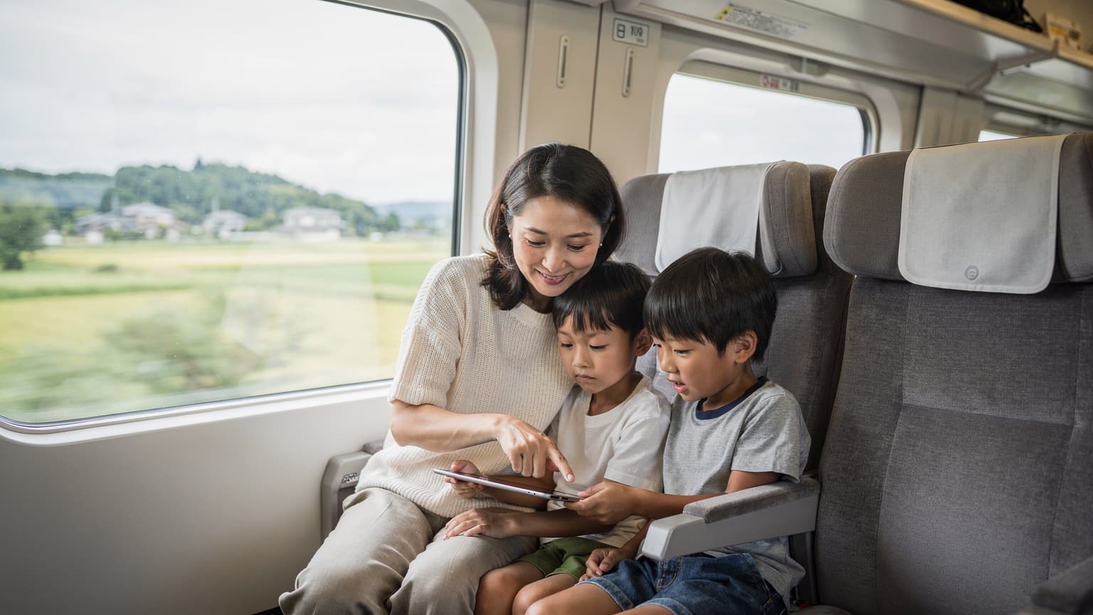 一位母親在東京地鐵新幹線車廂內,輕聲細語地指著平板電腦,兩個孩子專注地觀看,車窗外是模糊的日本鄉村景色。