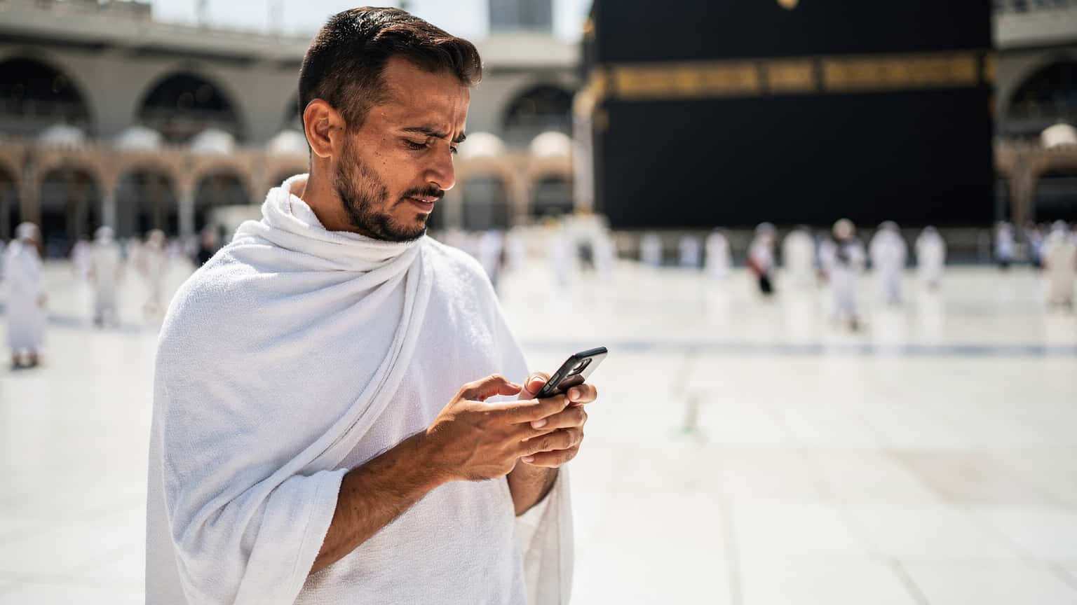 A pilgrim in traditional Ihram attire holding a smartphone, looking at a digital map application with the Kaaba visible in the background, under a clear blue sky in Mecca.