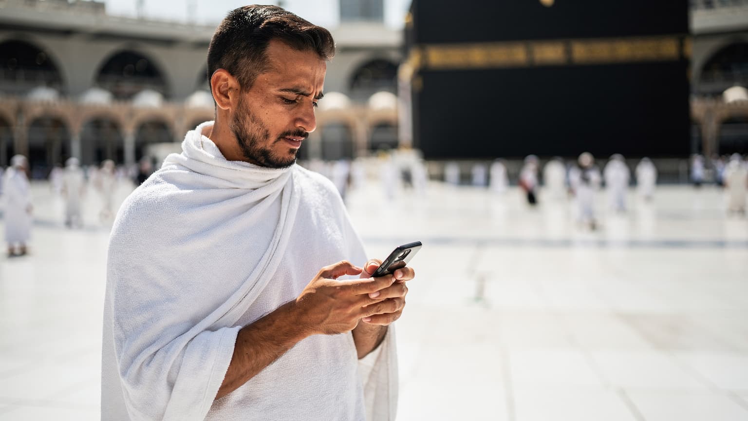 A pilgrim in traditional Ihram attire holding a smartphone, looking at a digital map application with the Kaaba visible in the background, under a clear blue sky in Mecca.