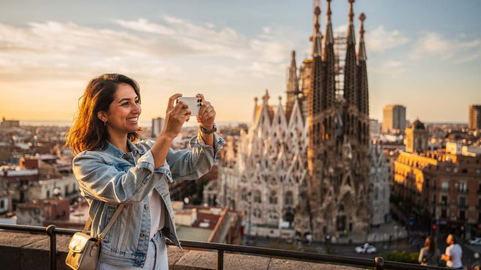 Una mujer sonriente, usando su iPhone para tomar una foto de la Sagrada Familia en Barcelona al atardecer, con la ciudad extendiéndose detrás de ella.