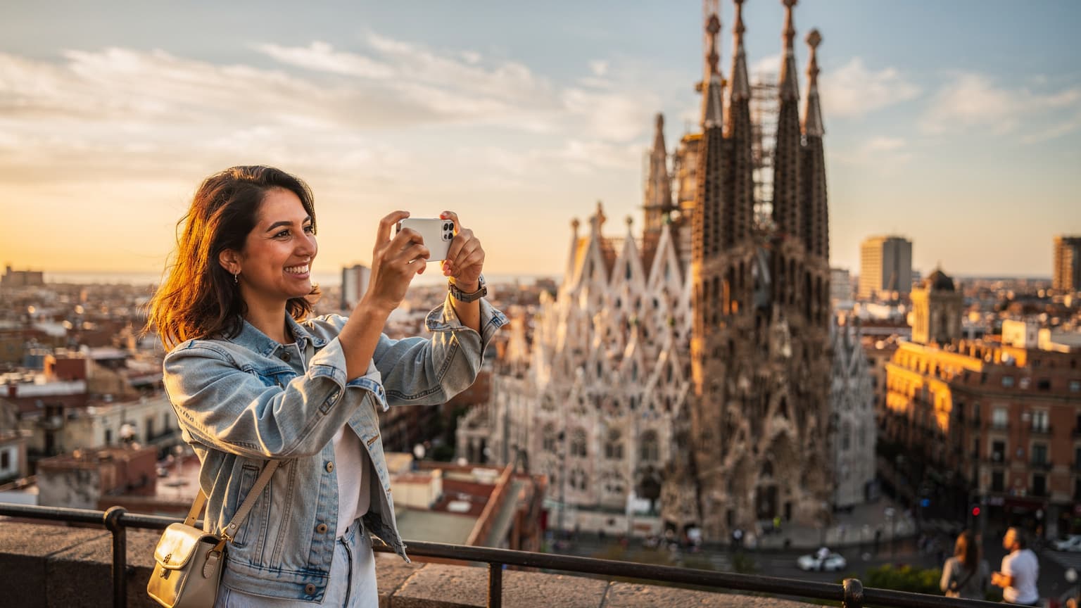 Una mujer sonriente, usando su iPhone para tomar una foto de la Sagrada Familia en Barcelona al atardecer, con la ciudad extendiéndose detrás de ella.