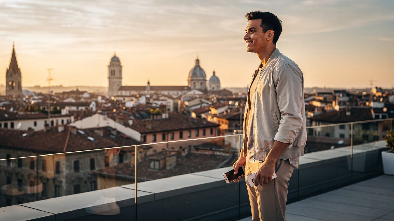 A photorealistic editorial image of a traveler overlooking a European city panorama at golden hour. The traveler holds a modern smartphone, symbolizing global connectivity and ease of travel. Iconic European architecture fills the background, bathed in warm light.