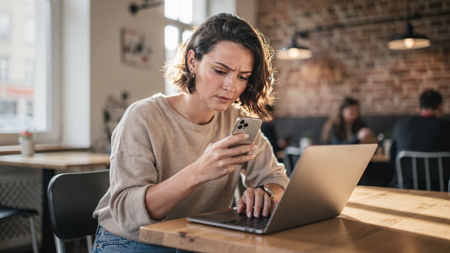 En kvinde sidder ved et bord på et travlt co-working space i Berlin, med sin iPhone i hånden og en kop kaffe ved siden af, mens hun arbejder fokuseret.
