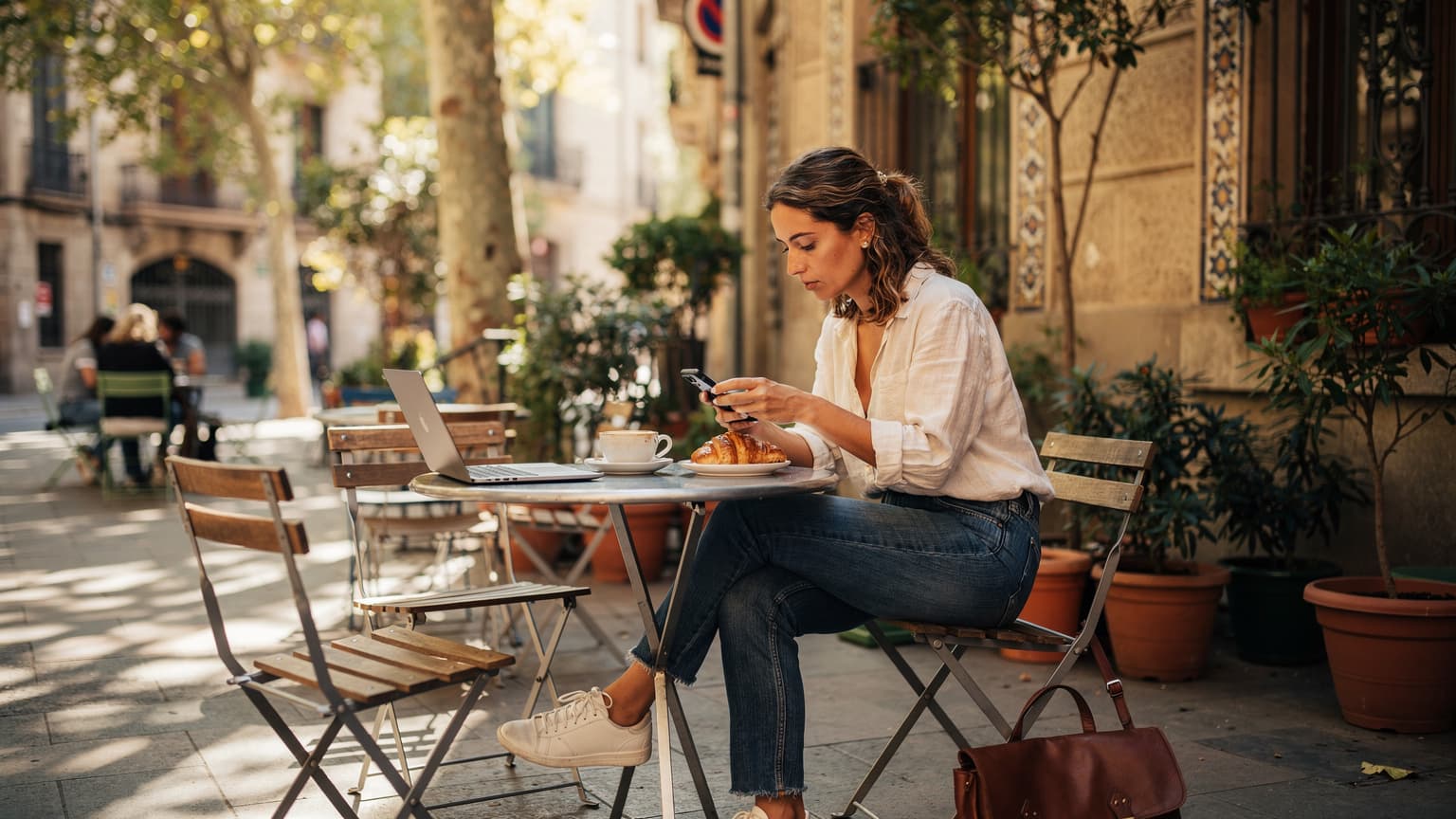 Una viajera sentada en la terraza de un café en el barrio de Gràcia, Barcelona, revisando su teléfono con una eSIM activa mientras disfruta de un café con leche y una tostada.
