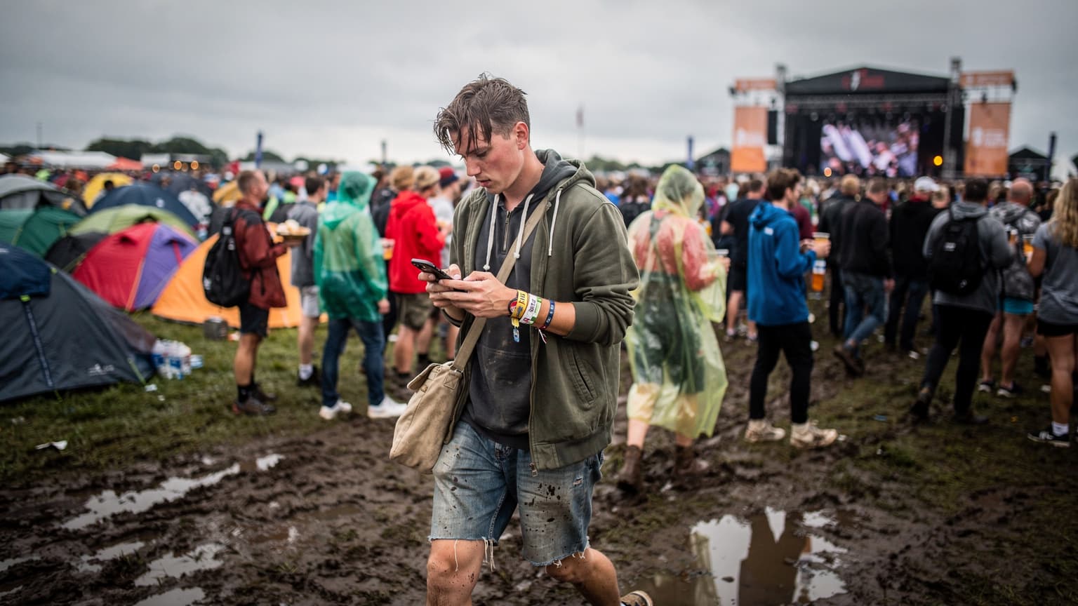 En ung festivalgænger med en smartphone i hånden navigerer gennem en tæt pakket menneskemængde på Roskilde Festival, hvor farverige telte kan ses i baggrunden.