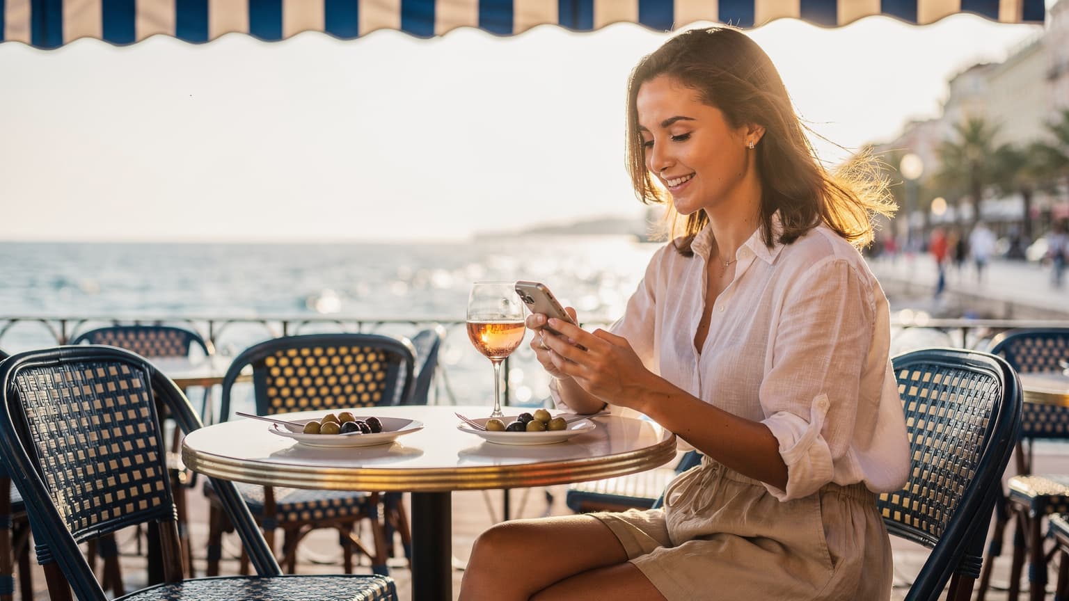 Une jeune femme souriante, assise sur la terrasse d'un café traditionnel à Nice, avec un smartphone à la main, un verre de rosé sur la table et la mer Méditerranée en arrière-plan sous un ciel bleu d'été.